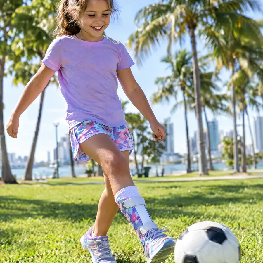 A smiling girl kicking a soccer ball while wearing a colorful ankle foot orthosis